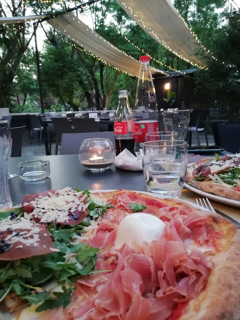 Due pizze servite in terrazza al tramonto, sotto i teli e le lucine, con il verde del giardino intorno